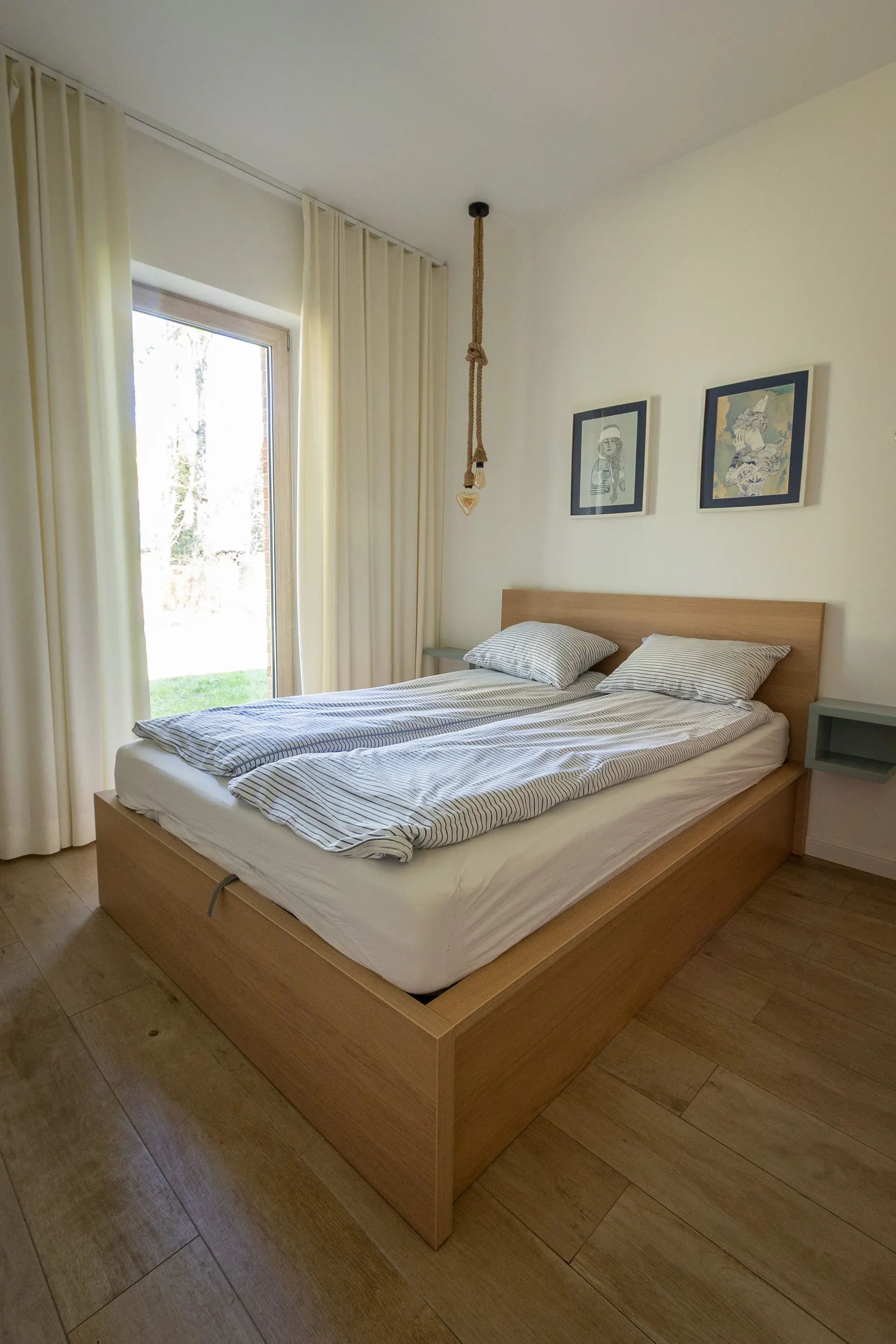 Sypialnia z podwójnym drewnianym łóżkiem i naturalnymi zasłonami przy dużym oknie z widokiem na ogród — Apartament 3 Poezja Ciszy Dębki